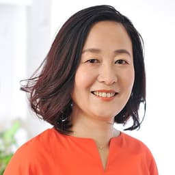 白い室内を背景に、うっすらとグリーンの植物が映り、全体的に明るく清潔感のある雰囲気。オレンジ色のトップスを着た女性のバストアップショット。体はやや右に傾け、顔は正面を向いて柔らかく微笑んでいる。髪は肩にかかる長さのゆるいカール。