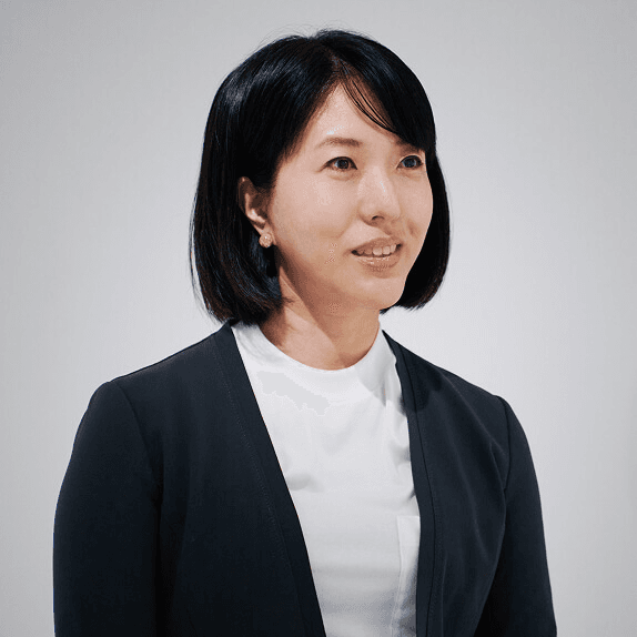 Bust-up shot of a woman against a white background. She is wearing a navy blue jacket and a white round-necked shirt, facing slightly to the right. Her hair is in a pigtail and parted 6:4 in the front.