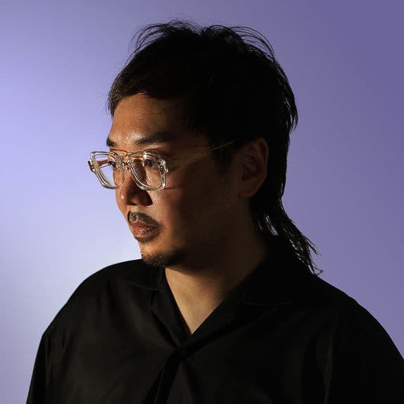 Bust-up shot of a man dressed in black against a purple background. He wears transparent glasses and is looking to the left. The light is strong from the left and casts a shadow on the back of the head area on the right side of the image.