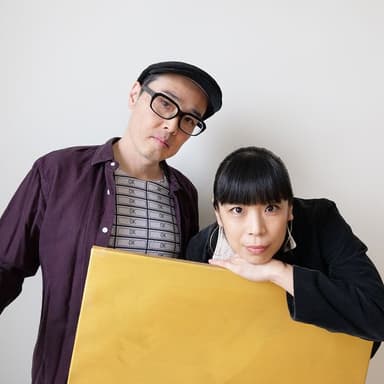 Two shots of a man and a woman lined up against a white background with a yellow board in front of them. The man on the left is wearing a purple shirt and a black hat, with black-rimmed glasses and his head tilted slightly toward the woman. The woman on the right is dressed in black, with her elbows on the board and her face close to the board, and her black hair is styled with a matching fringe. She wears large earrings.