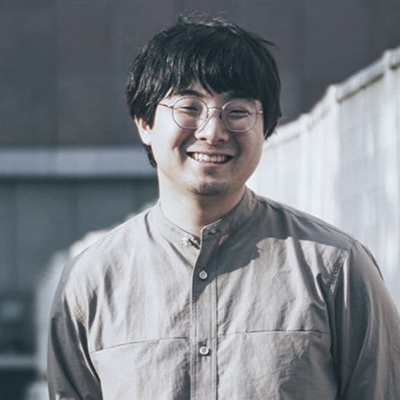 Bust-up shot of a man standing in front of a concrete-like background looking at us with a smile. He is wearing a collarless beige shirt and glasses.