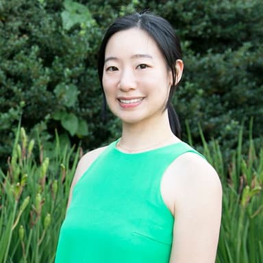 Bust-up shot of a woman wearing a bright green sleeveless one-piece dress against a background of tall trees, grass, and other green vegetation. Her black hair is pulled back in a single bun, and her body is turned slightly to the left while her face faces forward, smiling.