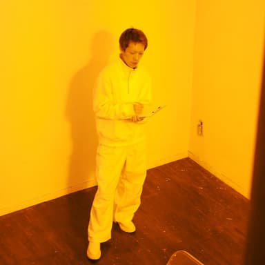 黄色くライティングされた空間に、白い服をきた短髪の男性が立っている